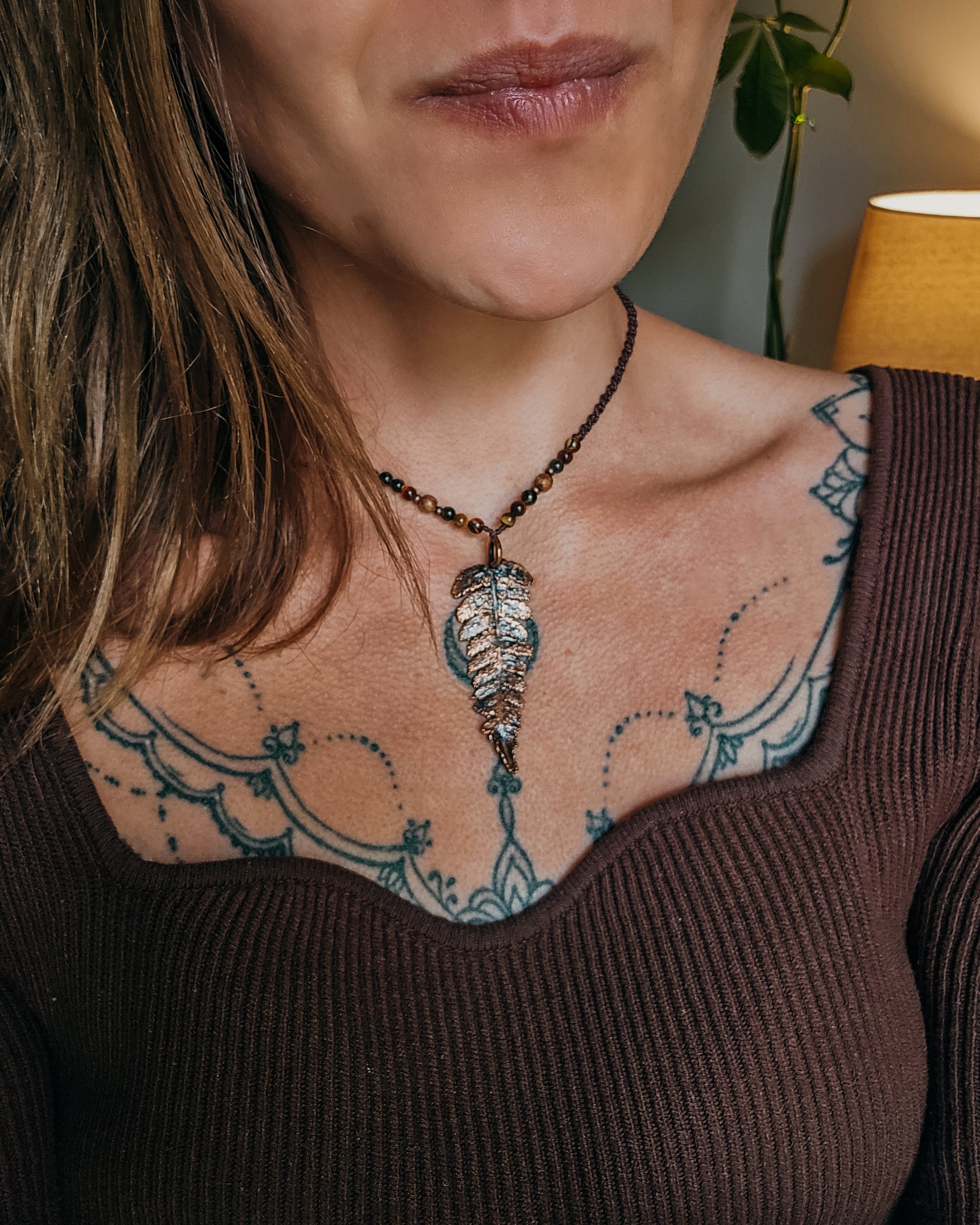 Close-up of a woman with tattoos on their shoulder and neck, wearing Necklace with tiger's eye and wooden beads and a copper electroformed fern charm. 