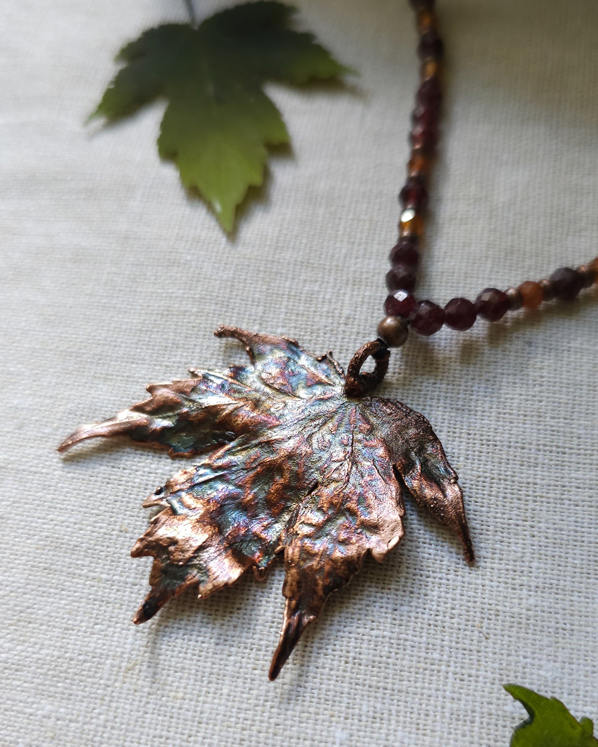 Handmade Copper electroformed Necklace with maple leaf pendant and Garnet crystal beads on a textured surface with leaves