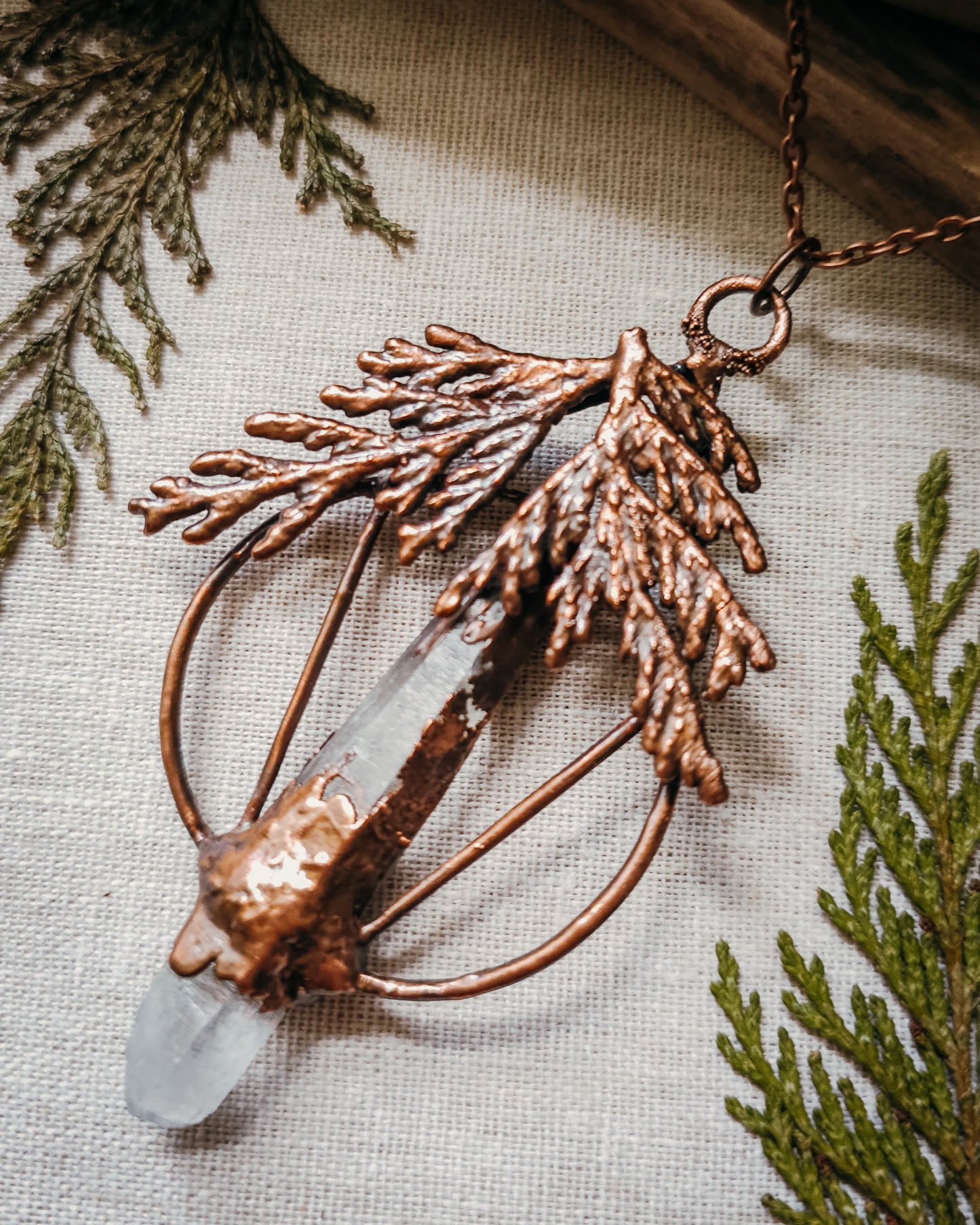 Handmade Copper electroformed Necklace with a cypress and clear Quartz crystal pendant on a textured fabric background