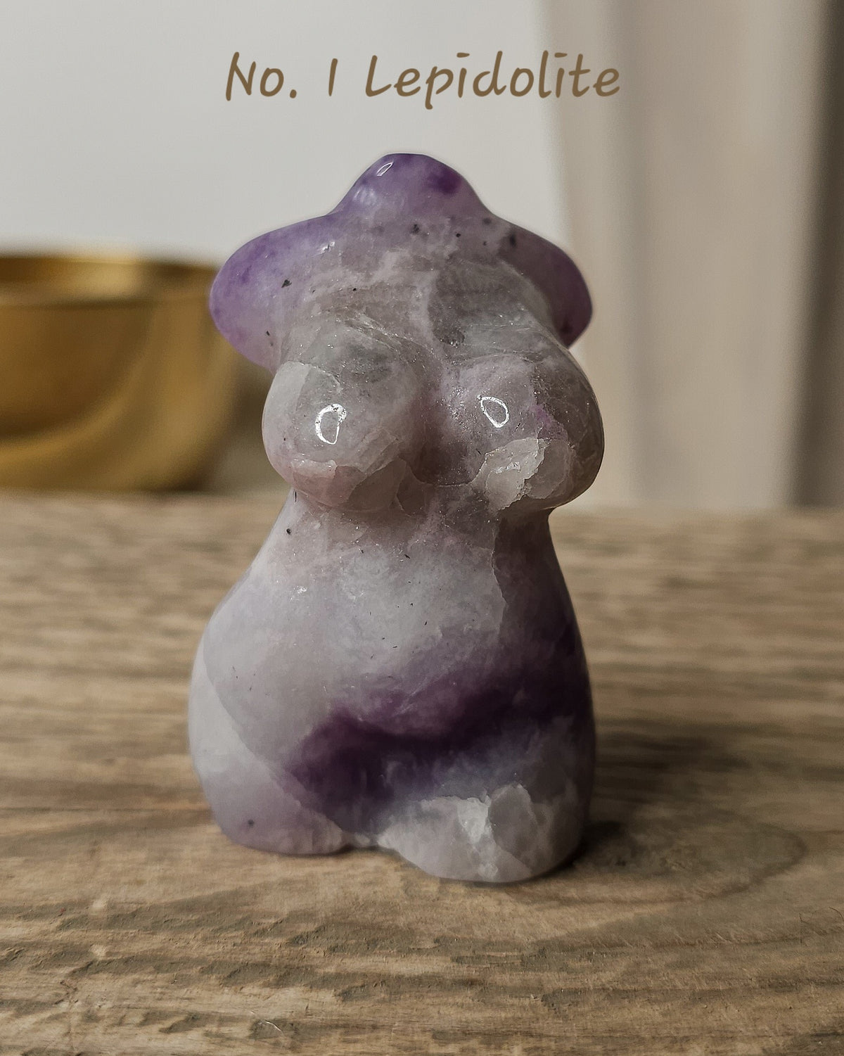 A small carved crystal figurine shaped like a  female form, made from lavender and purple lepidolite with pale gray-white areas. The figurine is placed on a wooden surface with a softly blurred indoor background