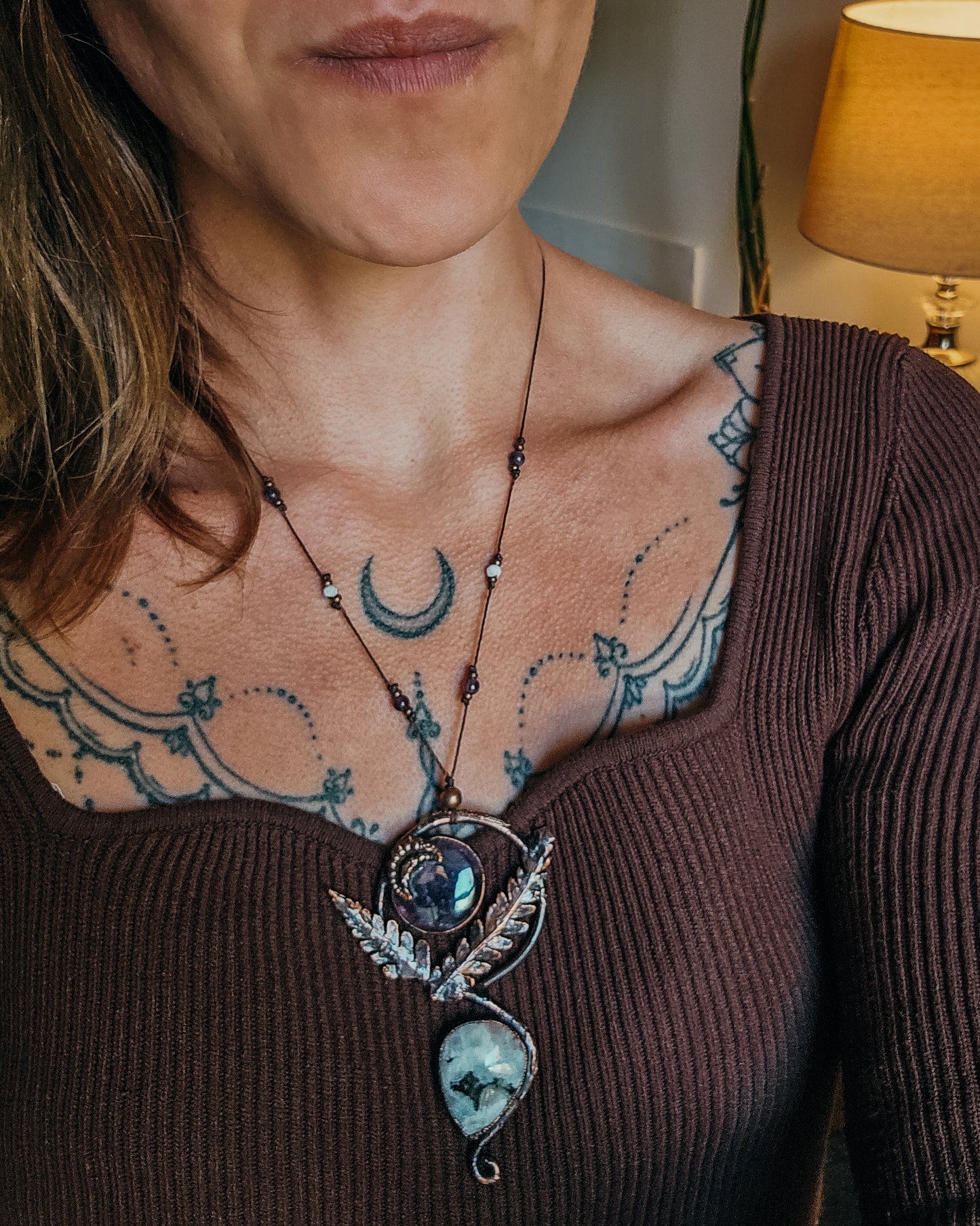 Close-up of a person wearing a  Copper electroformed Ornate pendant with amehtyst, Moonstone and fern leaves on a wooden surface with leaves with a brown top.