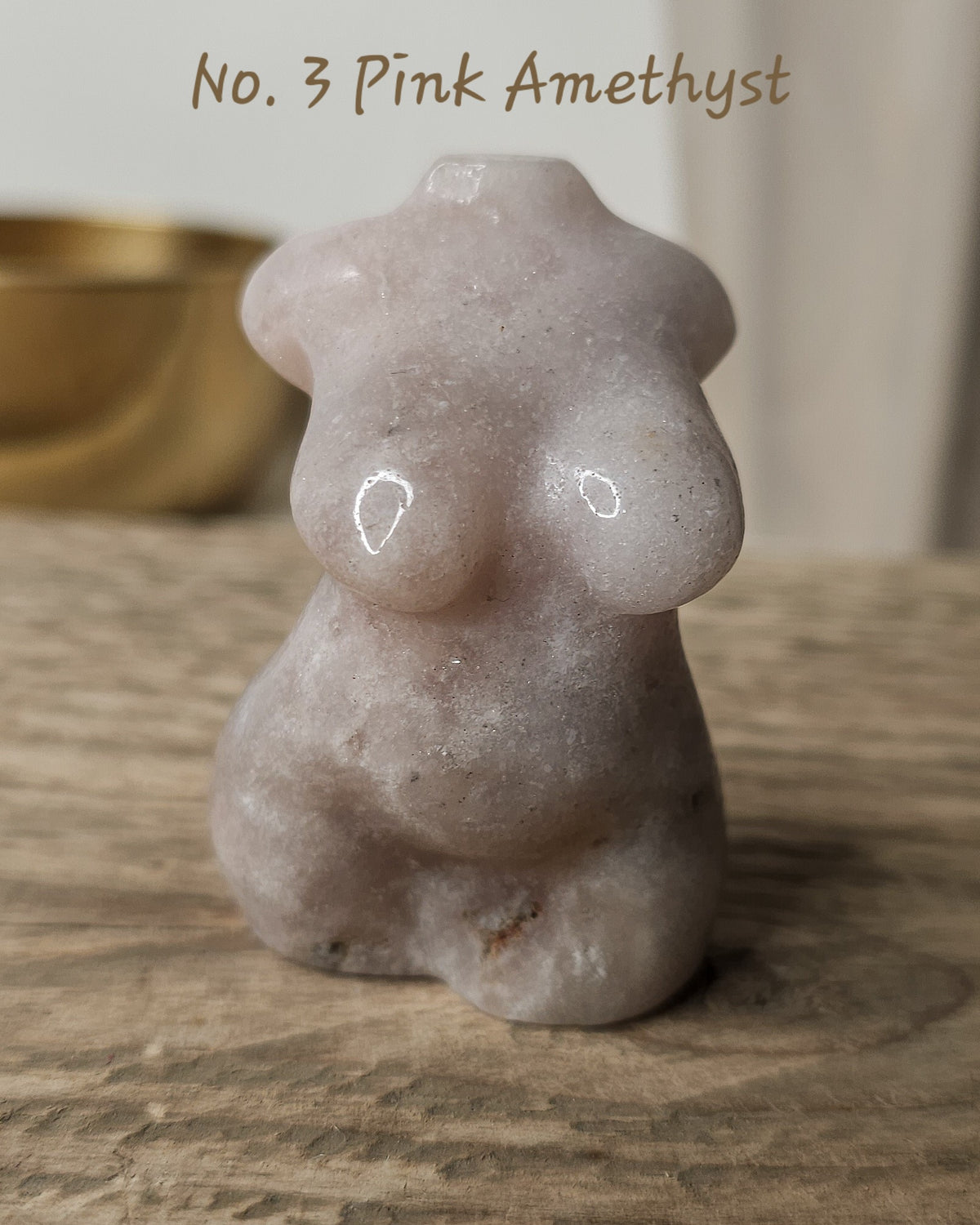 A small carved pink amethyst figurine shaped like a female body, displayed on a wooden surface with a blurred gold bowl in the background. 