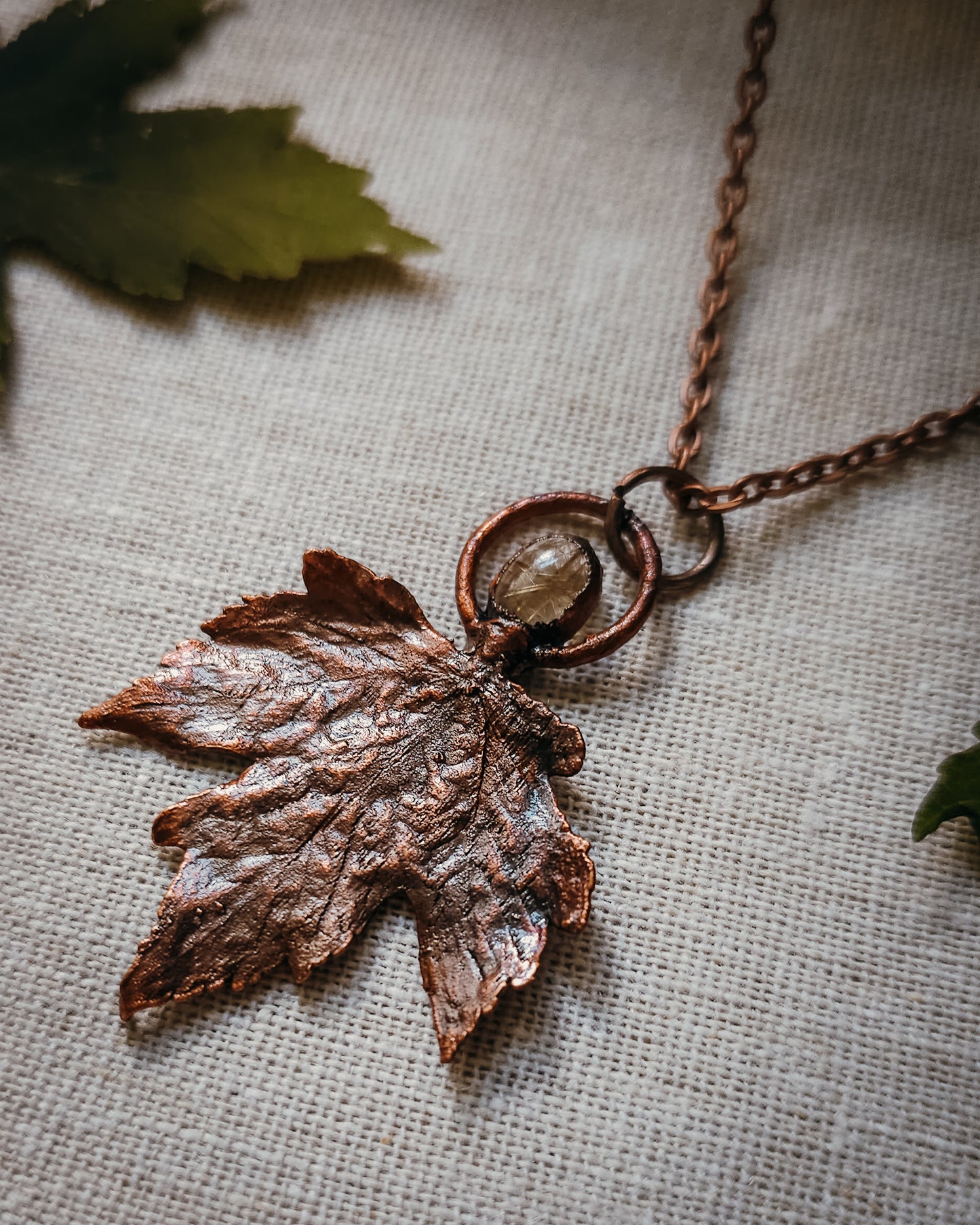 Copper electroformed Necklace with maple leaf pendant and golden rutilated Quartz crystal on a textured surface with leaves