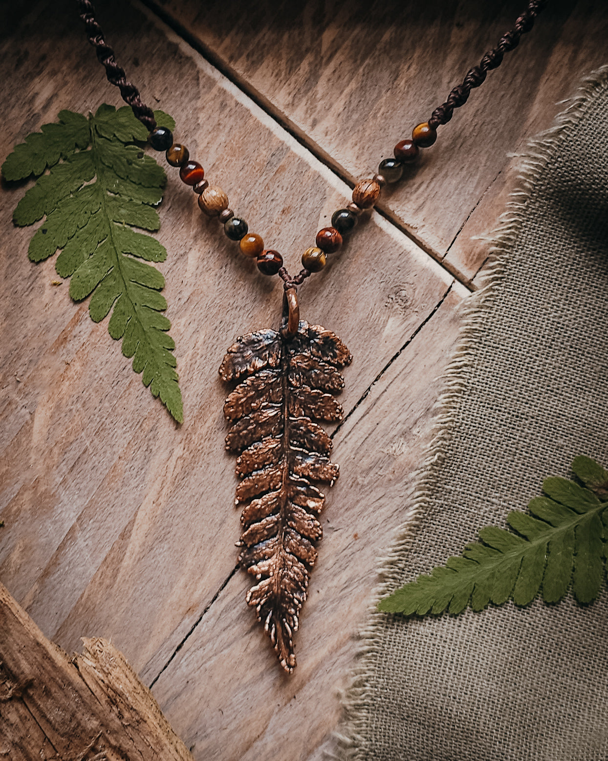 Necklace with  tiger's eye and wooden beads and a copper electroformed fern charm on a wooden surface with linen and fern leaf.