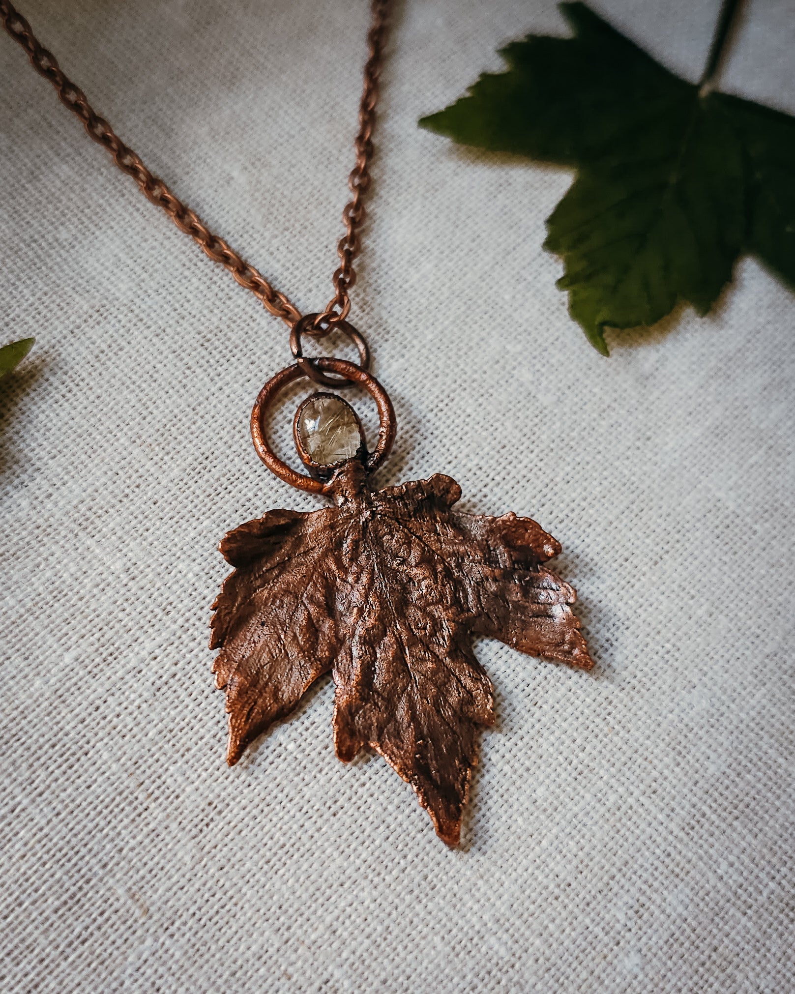 Copper electroformed Necklace with maple leaf pendant and golden rutilated Quartz crystal on a textured surface with leaves