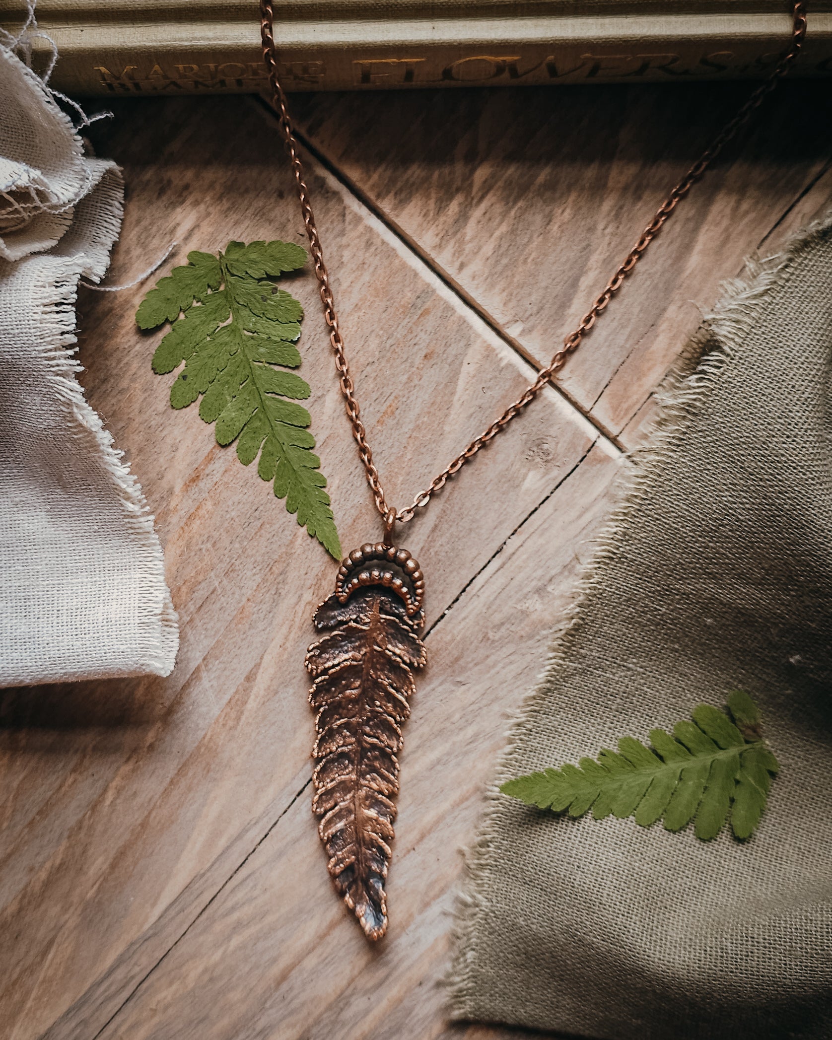 Copper electroformed fern pendant with crescent moon detail on a copper-coloured stainless steel chain, handcrafted from a real fern gathered in Scotland and photographed with fresh fern fronds on a rustic woodland-inspired background.