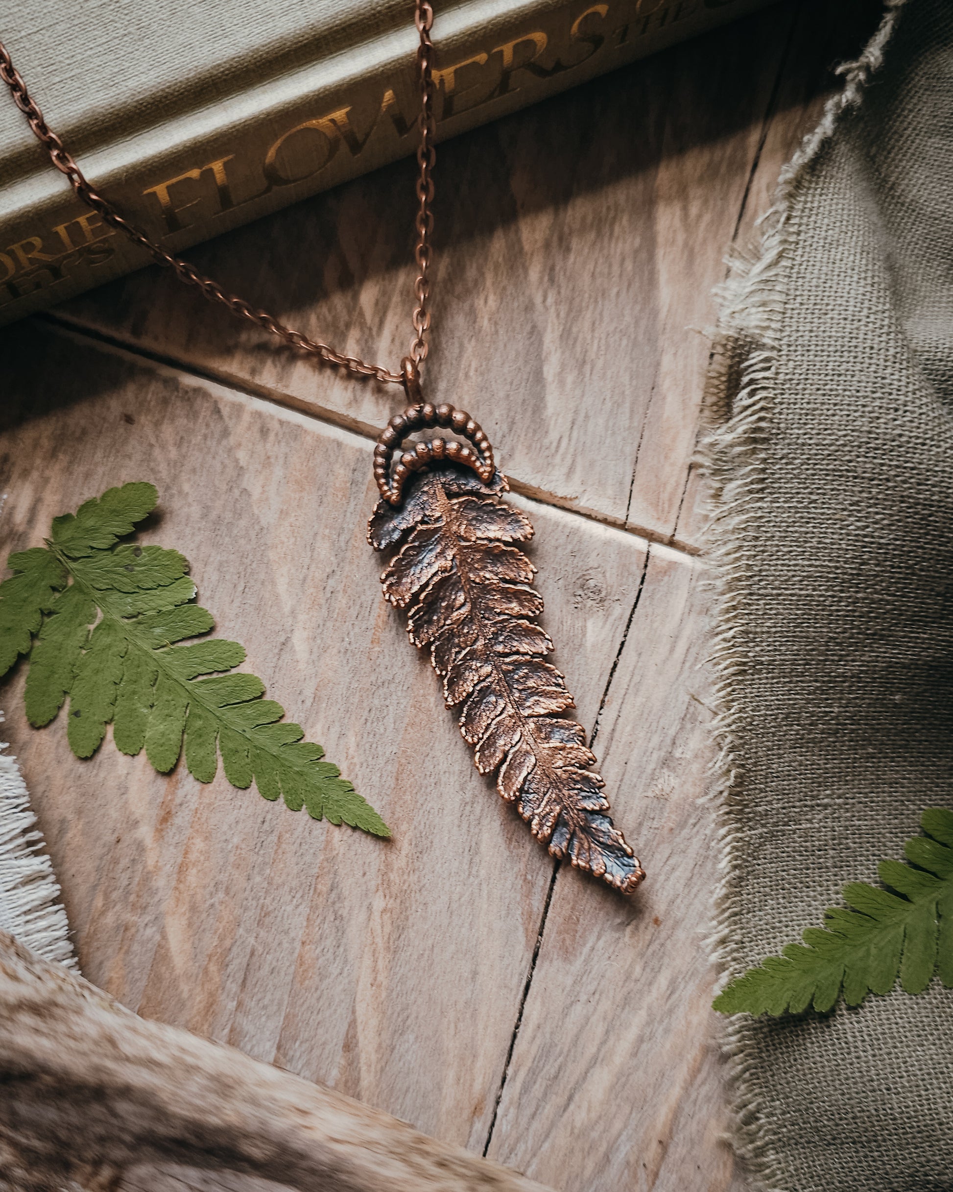 Copper electroformed fern pendant with crescent moon detail on a copper-coloured stainless steel chain, handcrafted from a real fern gathered in Scotland and photographed with fresh fern fronds on a rustic woodland-inspired background.