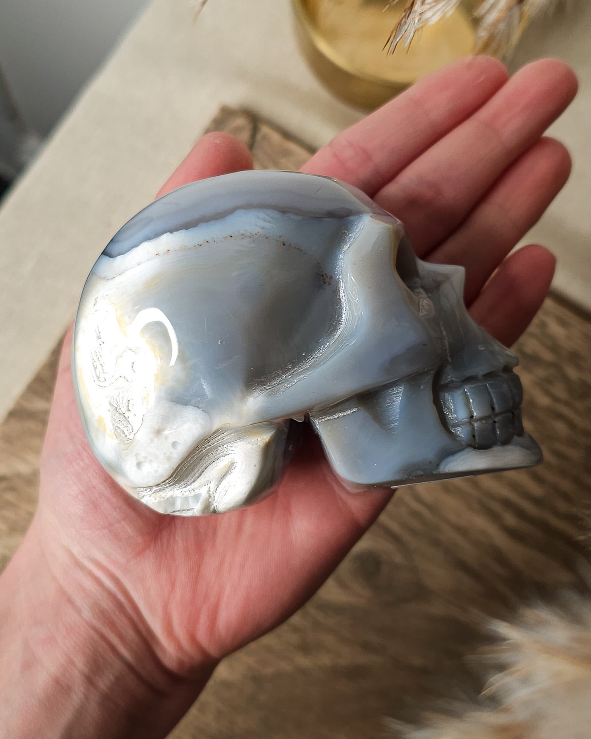 Blue Agate skull carving with layered pale blue and grey bands, a natural brown inclusion along the side and a sparkling druzy crystal cavity at the top.