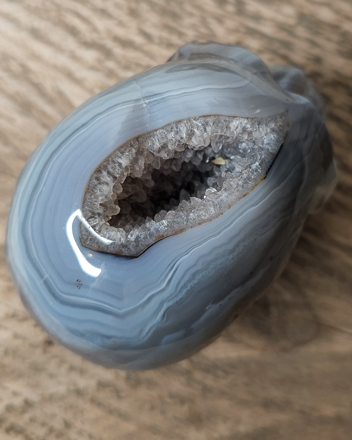 Blue Agate skull carving with layered pale blue and grey bands, a natural brown inclusion along the side and a sparkling druzy crystal cavity at the top.